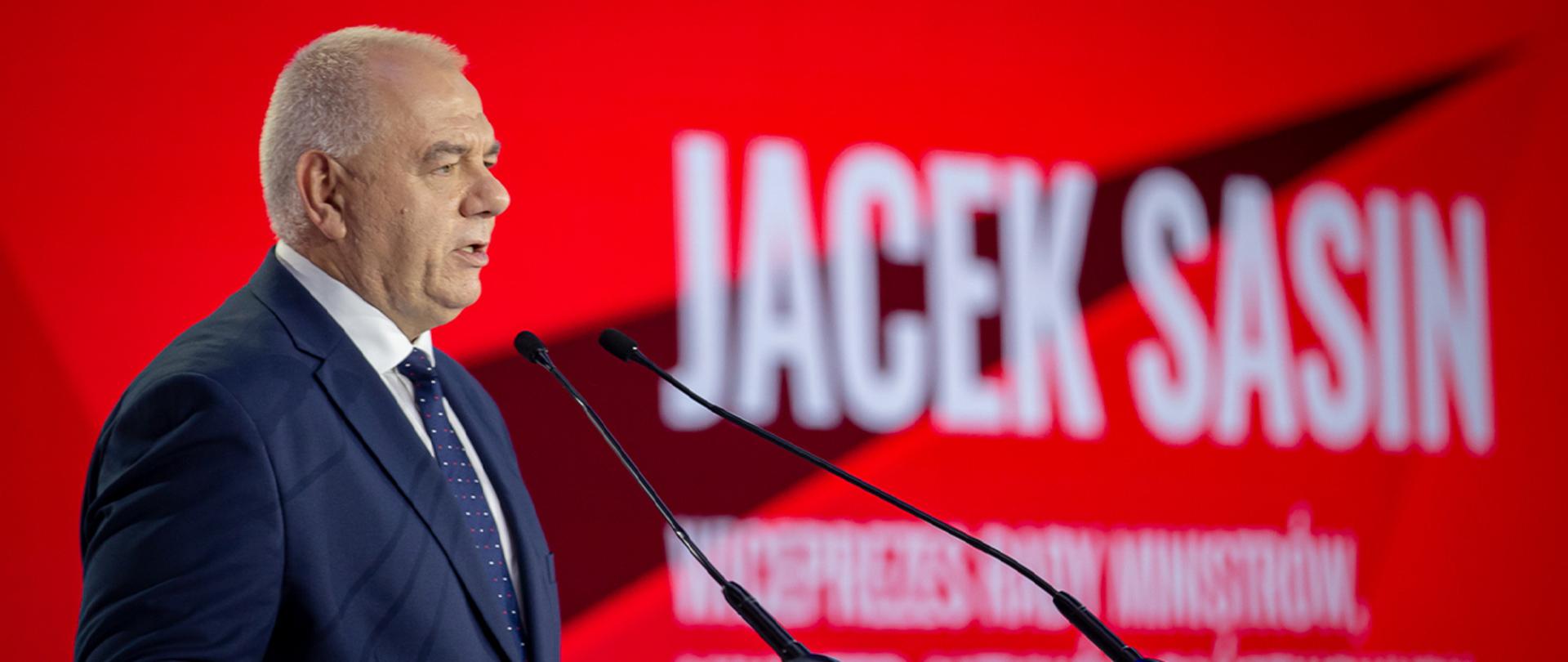 Wicepremier Jacek Sasin przemawia podczas Kongresu 590 przy mikrofonie. W tle biały napis na czerwonym tle: Jacek Sasin, Wiceprezes Rady Ministrów.
