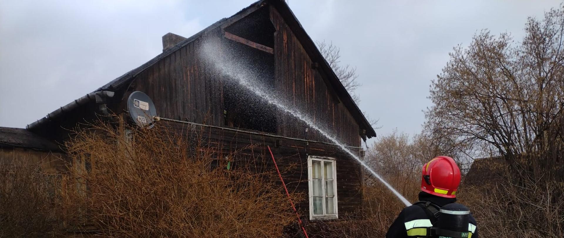 Na zdjęciu widać rozłożone węże strażackie przed budynkiem, w którym
doszło do pożaru. Na drugim planie stoją dwa samochody pożarnicze na drodze.