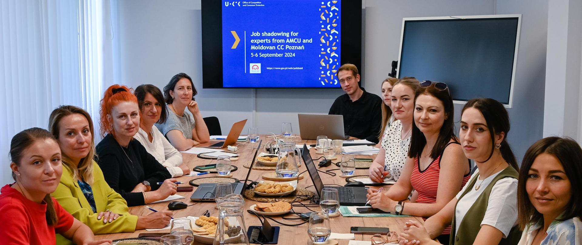 W sali szkoleniowej przy stole konferencyjnym 11 osób z laptopami oraz ciasteczkami. Z tyłu wyświetlana jest prezentacja Job shadowing for experts from AMCU and Moldovan CC Poznań z logo Polska pomoc
