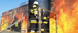 Komora ogniowo rozgorzeniowa na niej namalowane płomienie oraz trzech strażaków. Z otwartych drzwi komory wydobywa się dym i dwóch strażaków jeden wychodzi a drugi stoi tyłem.