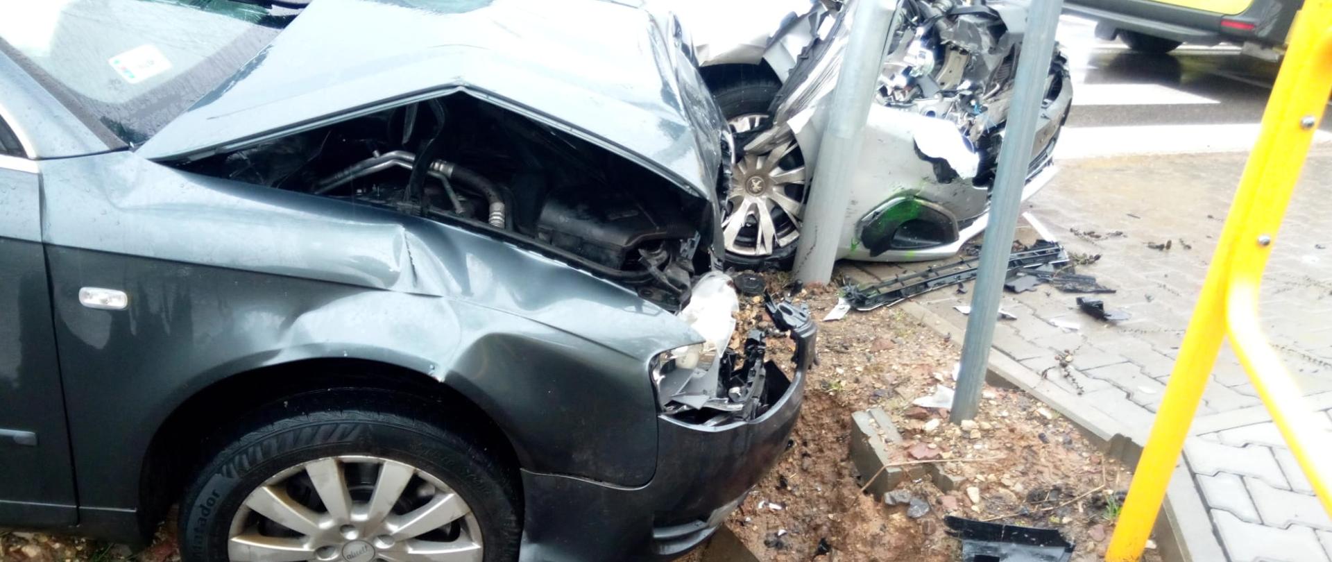 Zdjęcie przedstawia przody dwóch samochodów osobowych zniszczone w wyniku zderzenia. Stoją na poboczu, a jeden opiera się o latarnię uliczną. 