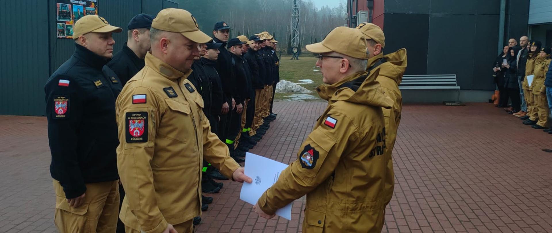 Przejście na zaopatrzenie emerytalne aspiranta Tomasza Wojdala. Komendant Powiatowy Państwowej Straży Pożarnej w Brzezinach starszy brygadier Mariusz Kaczmarski wręcza decyzję zwalniającą funkcjonariusza ze służby. Podczas wręczania decyzji asystuje zastępca Komendanta Powiatowego Państwowej Straży Pożarnej w Brzezinach młodszy brygadier Jakub Brzózka. Sytuacja ma miejsce podczas uroczystej zbiórki na placu komendy, strażacy stoją w dwóch szeregach na przeciw siebie.