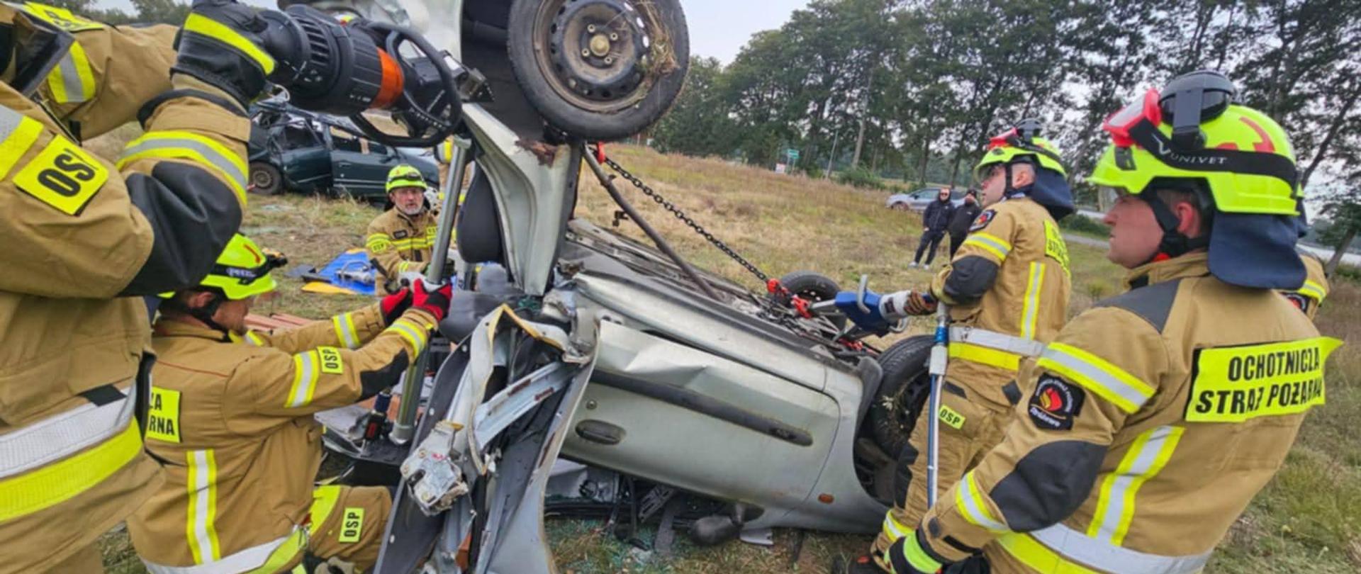 Zdjęcie przedstawia strażaków podczas ćwiczeń.
W tle samochody strażackie i tereny zielone.
