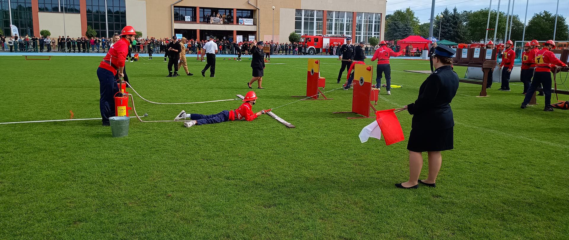 Na zdjęciu widoczna jest drużyna młodzieżowa w trakcie wykonywania ćwiczenia bojowego. W tle widać sędziów zawodów oraz pozostałych uczestników zawodów i zaproszonych gości. W oddali widoczny budynek szkoły.