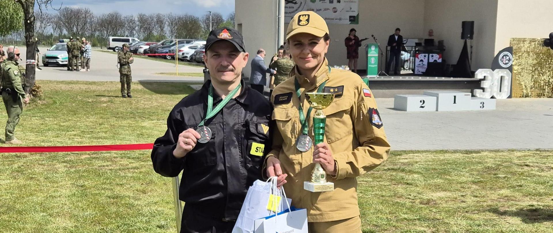 Na zdjęciu stoi dwóch strażaków. Mężczyzna w czarnym mundurze i kobieta w mundurze piaskowym. Mężczyzna trzyma w ręku medal, kobieta puchar.