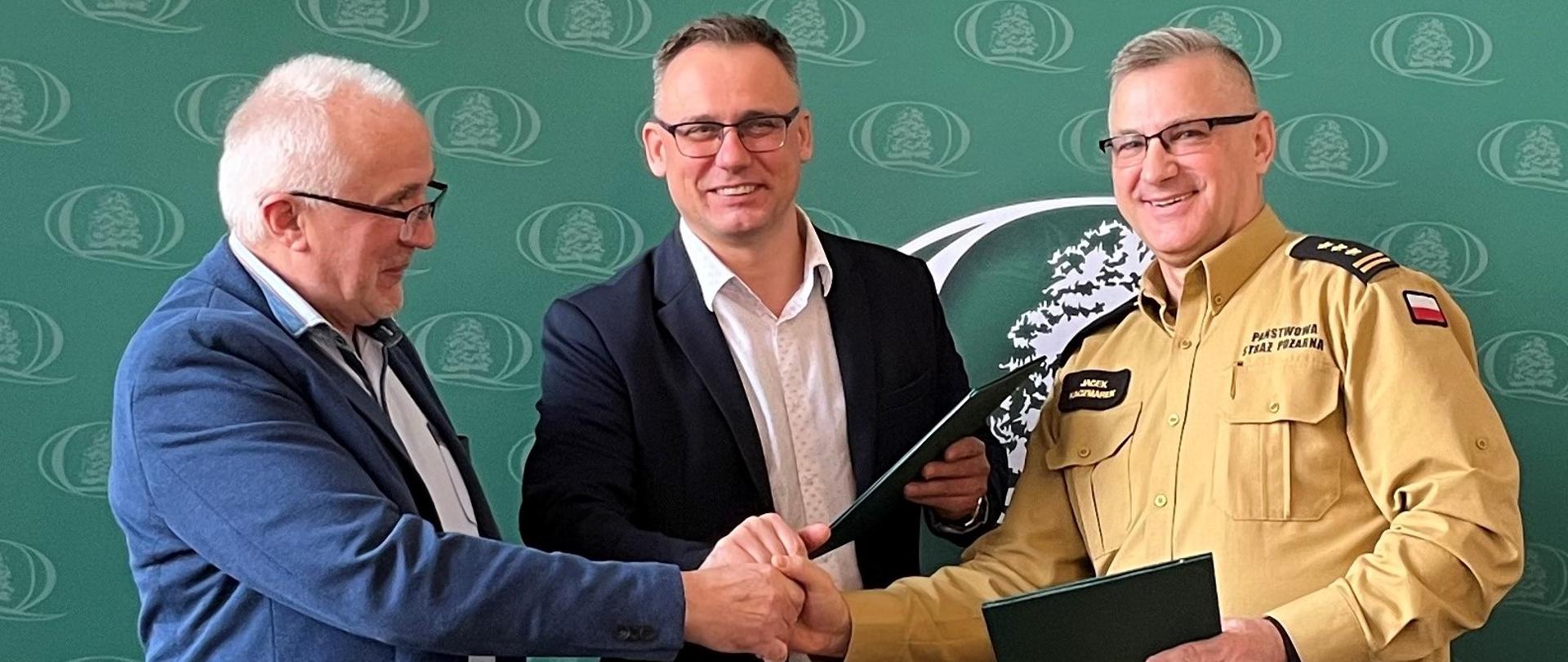 Przedstawiciele Wojewódzkiego Funduszu Ochrony Środowiska oraz kujawsko-pomorski komendant wojewódzki wymieniają dokumenty podpisanej miedzy sobą umowy. W tle ścianka z logo WFOŚiGW.