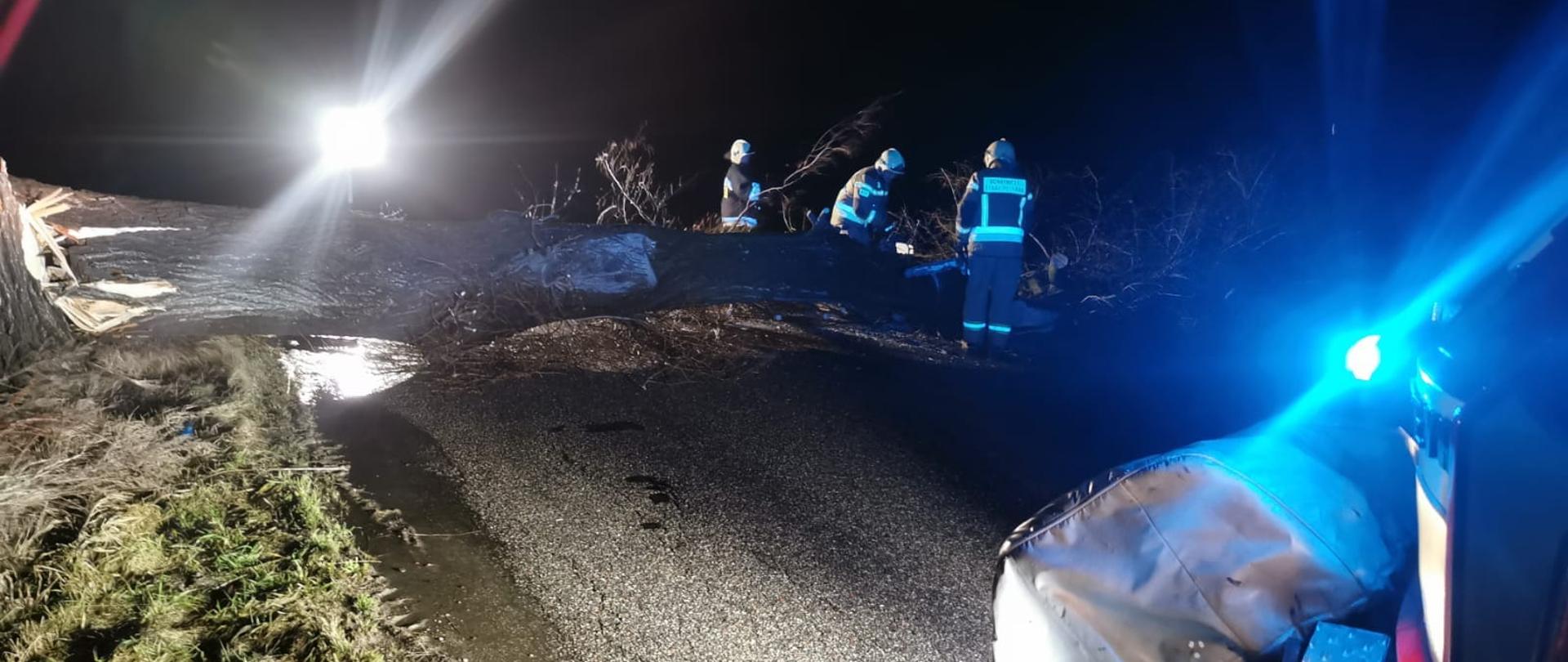 Pora nocna, na drodze w poprzek leży wielki konar drzewa który blokuje ruch w obu kierunkach. Trzech strażaków w ubraniach specjalnych koloru piaskowego i białych hełmach przy [pomocy piły spalinowej usuwają drzewo z pasa ruchu.