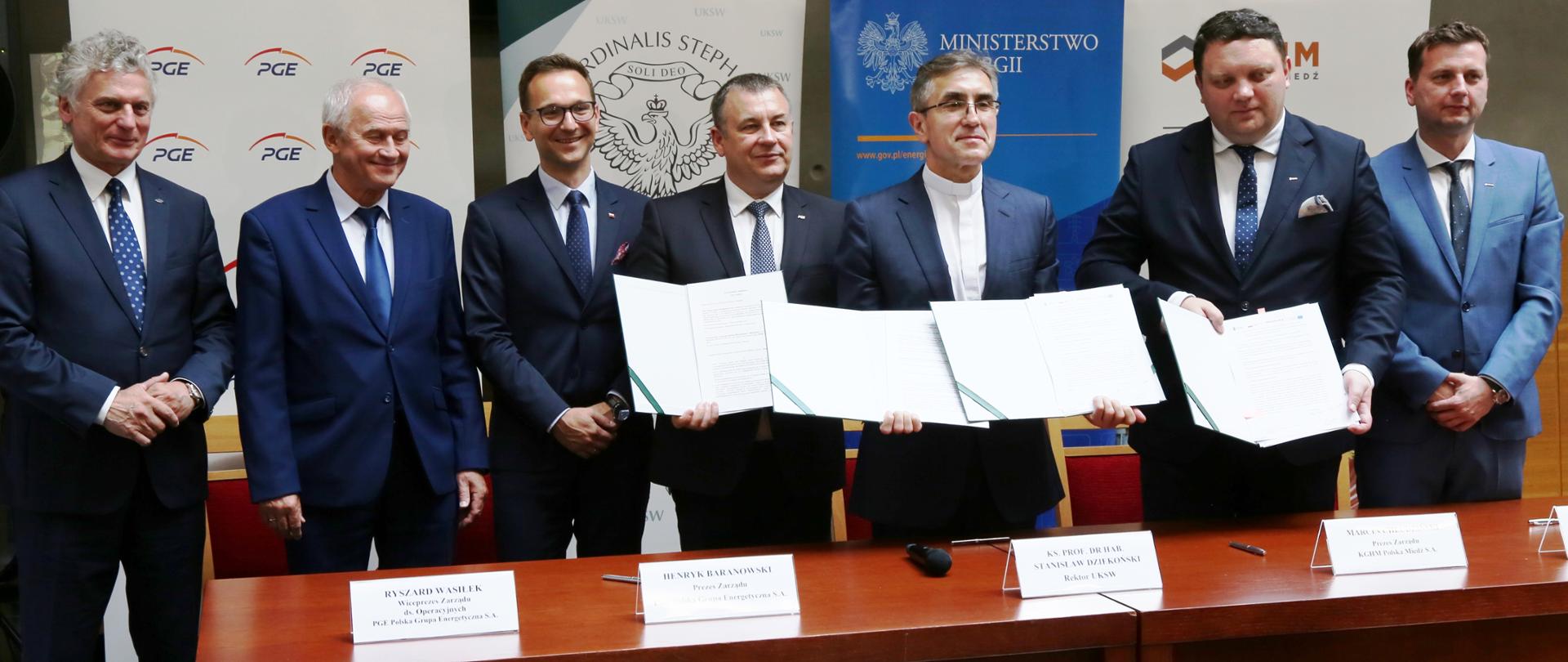 Uczestnicy uroczystości stoją za stołem i trzymają przed sobą podpisane porozumienia