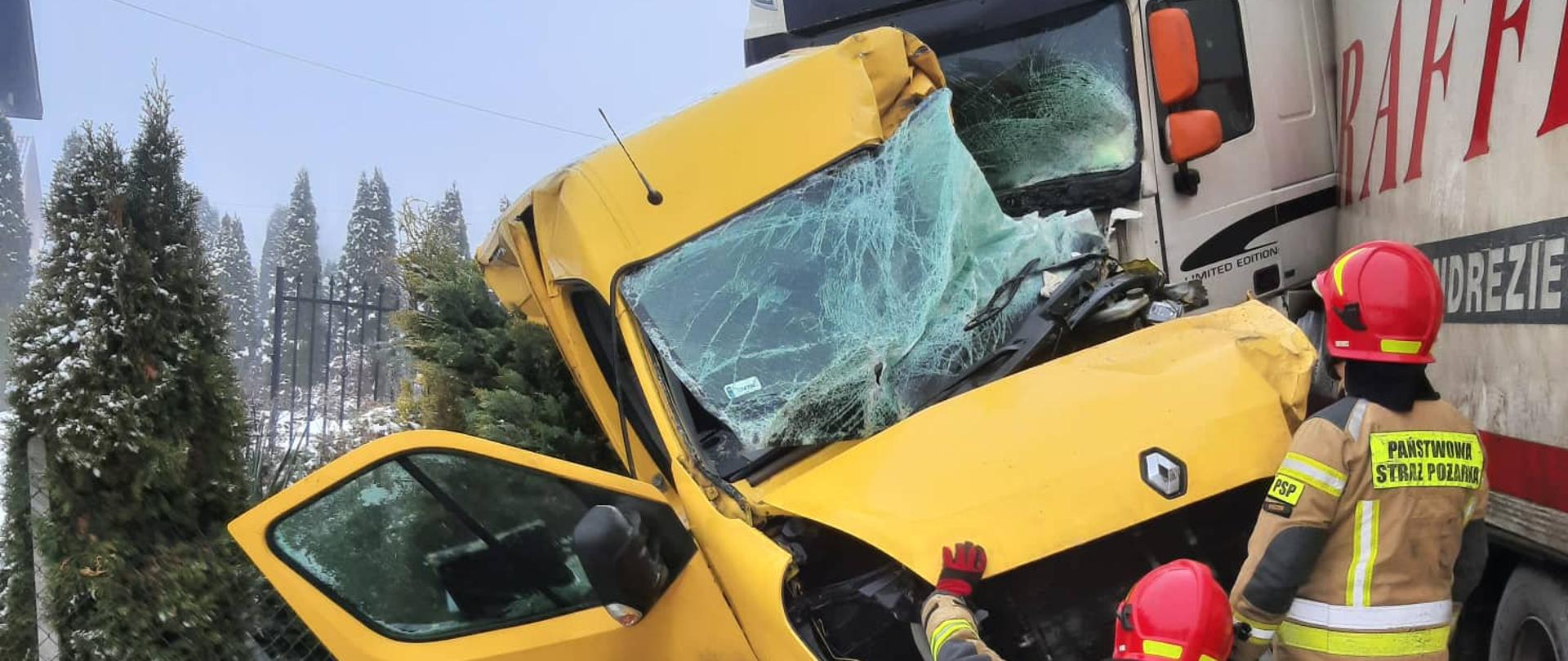 Na zdjęciu dwaj strażacy prowadzą działania w obrębie dwóch uszkodzonych pojazdów: dostawczego busa oraz ciągnika siodłowego z naczepą. 