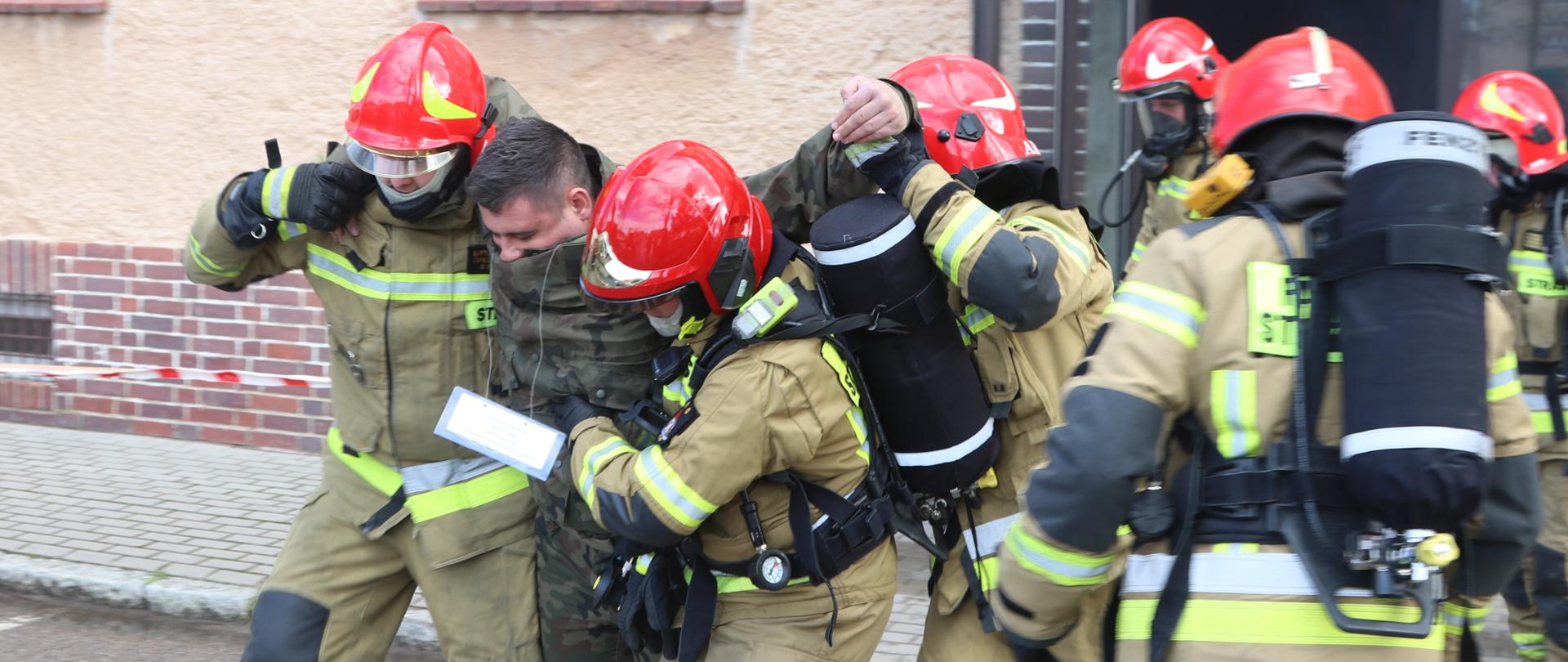 Strażacy w ubraniu specjalnym i zabezpieczeni w OUO ewakuują osobę poszkodowaną ze strefy zagrożenia. 