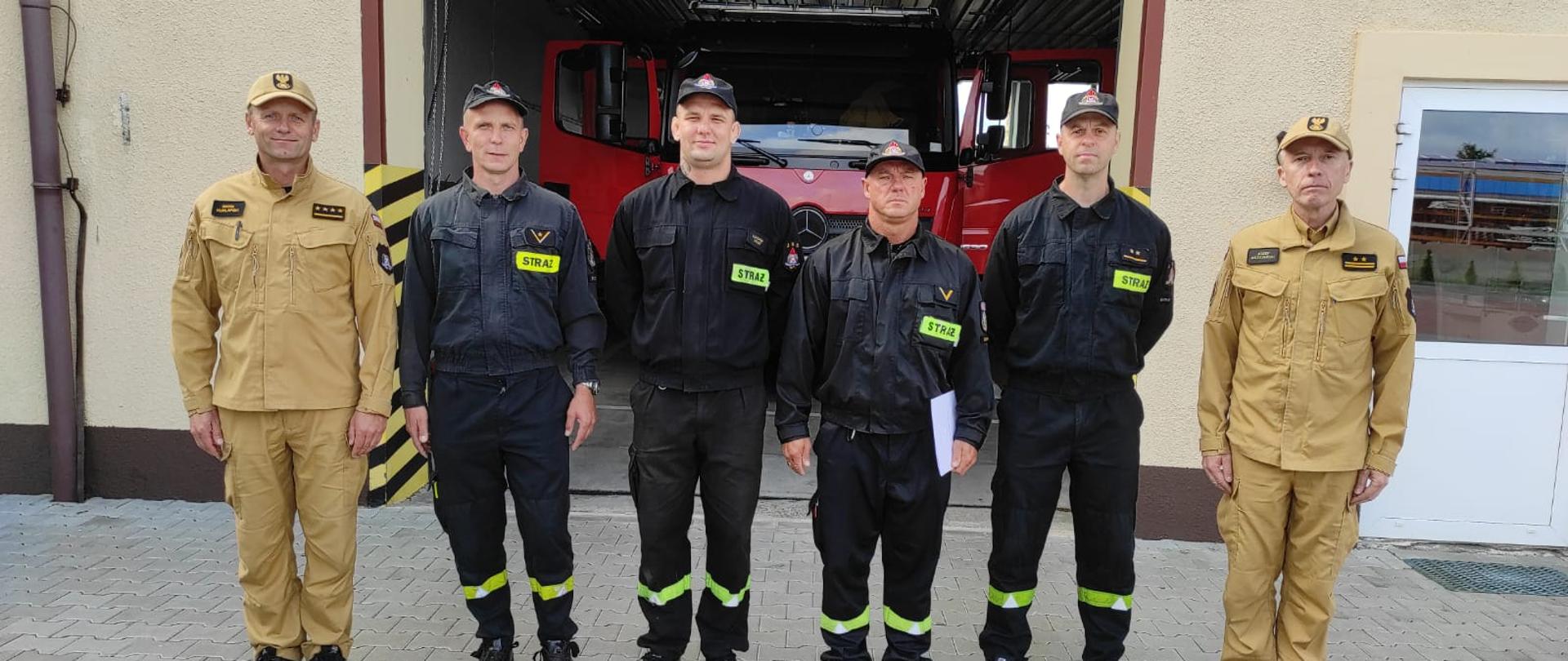 Zdjęcie przedstawia 6 strażaków stojących przed brama garażową pozując do zdjęcia. Za nimi pojazd pożarniczy w garażu.