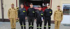 Zdjęcie przedstawia 6 strażaków stojących przed brama garażową pozując do zdjęcia. Za nimi pojazd pożarniczy w garażu.