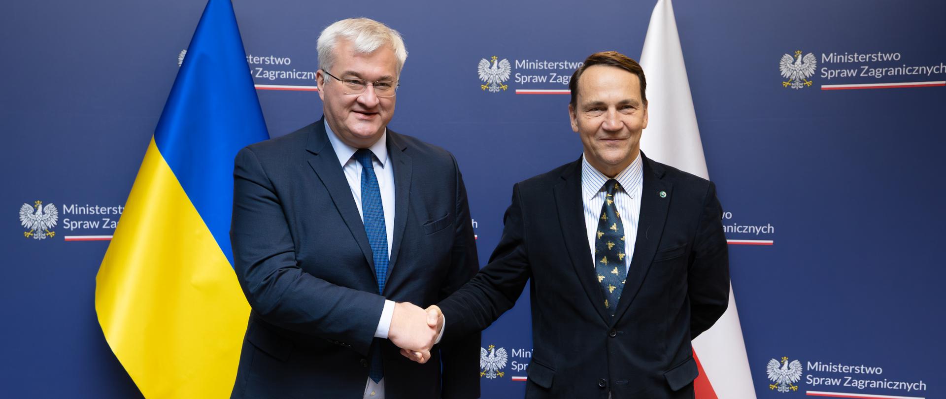 Na zdjęciu jest dwóch mężczyzn w garniturach. Podają sobie dłonie. Patrzą w stronę obiektywu. Za nimi jest granatowa ścianka z napisem "Ministerstwo Spraw Zagranicznych" i dwie flagi - Polski i Ukrainy