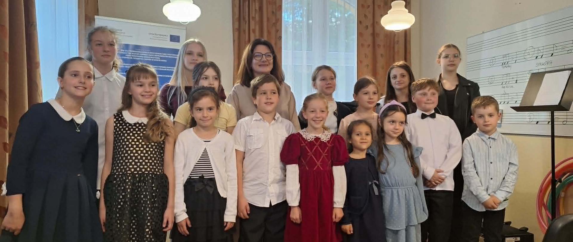 Uczestnicy: W centrum uwagi znajduje się grupa uśmiechniętych dzieci w różnym wieku, ubranych w eleganckie, galowe stroje (sukienki, koszule). W tle, za dziećmi, stoją starsze dziewczęta oraz dorosła kobieta w okularach. Otoczenie: Po prawej stronie widoczna jest biała tablica z narysowaną pięciolinią i zapisanymi nutami, a obok niej stoi pulpit na nuty. W tle po lewej stronie widać plakat z logotypem Unii Europejskiej, a u góry wiszą klasyczne, okrągłe lampy.