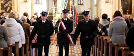 Na zdjęciu trzej strażacy w umundurowaniu służbowym, w czapkach rogatywkach, jeden ze strażaków trzyma sztandar. Idą przez środek kościoła, w ławkach zgromadzeni wierni, delegacje różnych instytucji i organizacji.