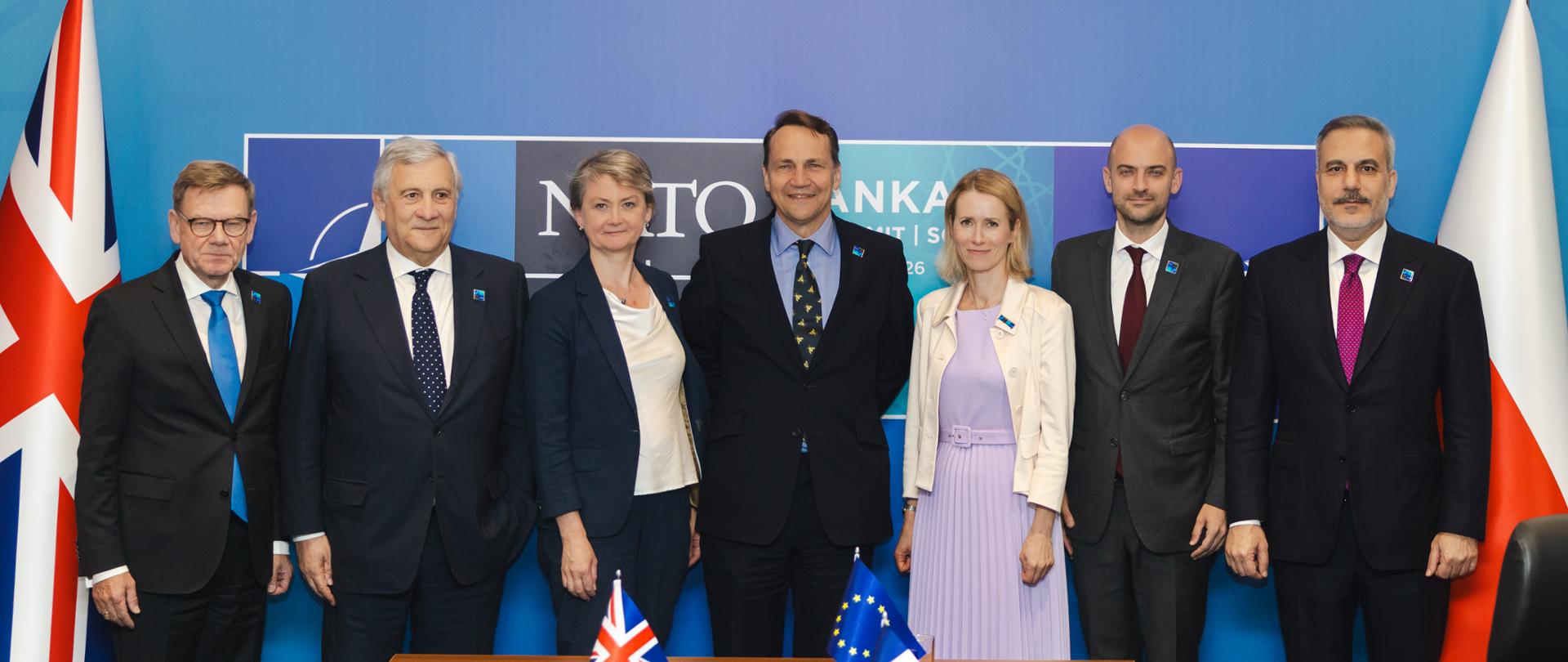 Participants of E5+ format meeting are posing for a group photograph. Deputy Prime Minister Sikorski is standing in the centre. In front of the group is a conference table with small flags of the United Kingdom, France and the European Union, bottles of water, glasses and documents. In the background, the flags of the United Kingdom and Poland can be seen, along with a blue backdrop featuring the NATO summit logo.