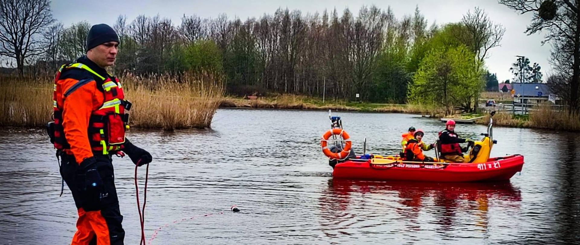 Brzeg jeziora. Z lewej strony strażak w kombinezonie i kamizelce do pracy w wodzie. Dalej na wodzie łódź strażacka i trzech strażaków na łodzi.