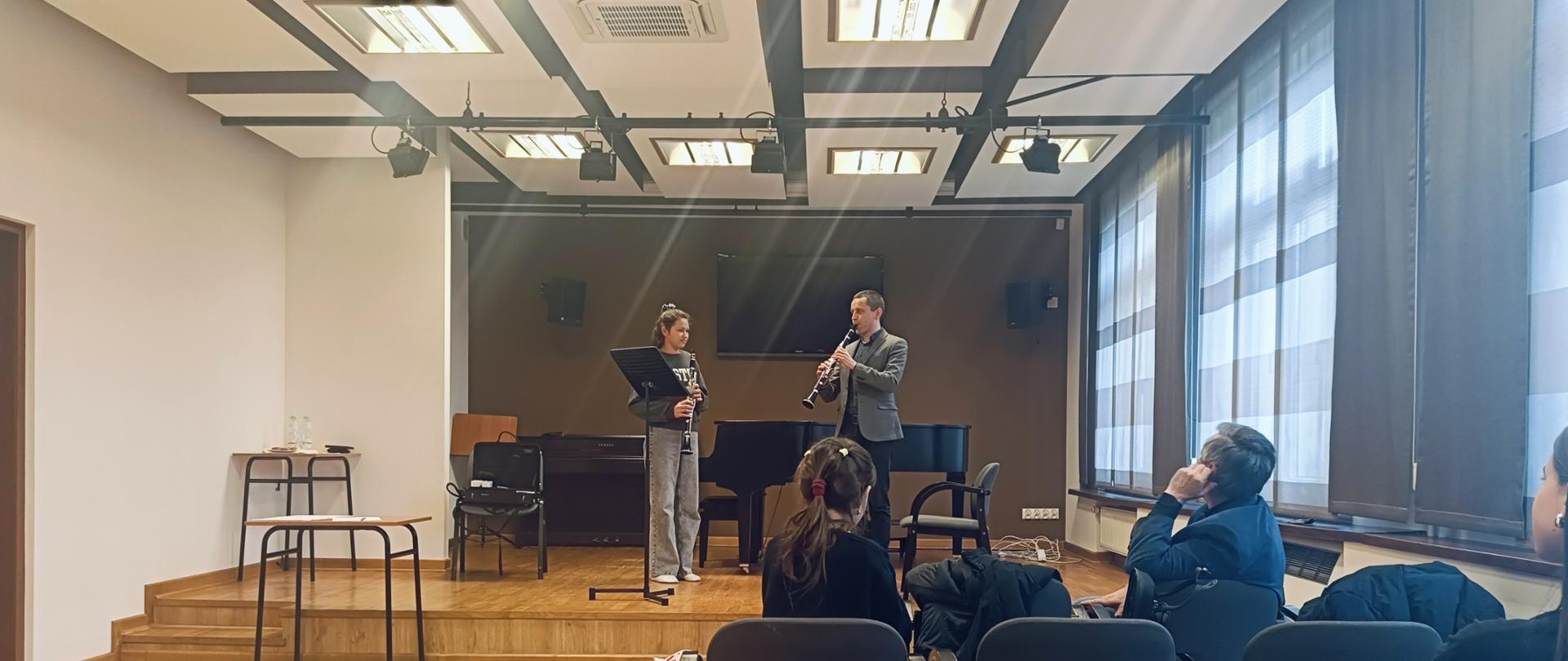 Wykładowca oraz uczestnik z klarnetami stojący na scenie przed pulpitem - prowadząćy wykonuje ćwiczenia