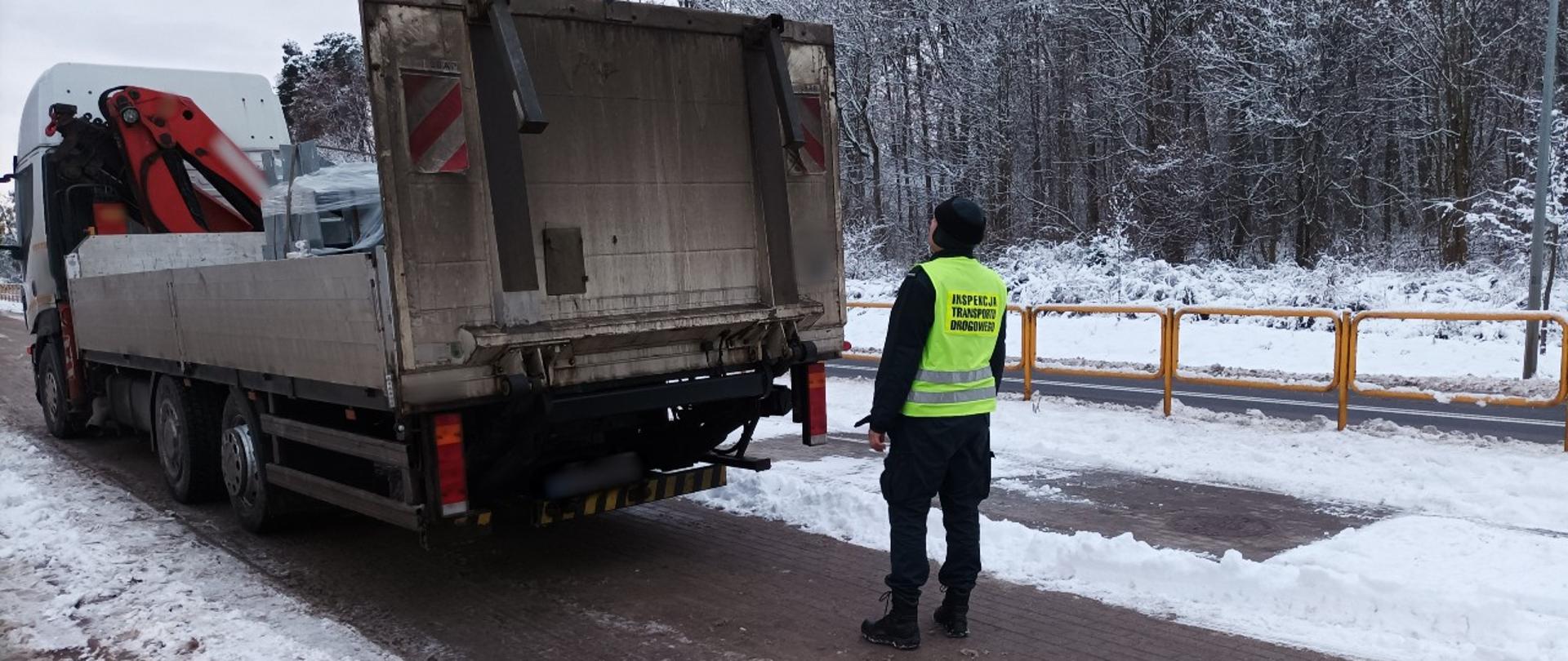 Umundurowany funkcjonariusz warmińsko-mazurskiej Inspekcji Transportu Drogowego stoi za kontrolowanym samochodem ciężarowym. Pojazd jest załadowany materiałami budowlanymi.
