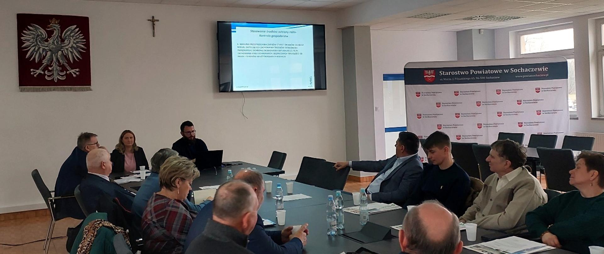 Uczestnicy spotkania siedzą przy stole konferencyjnym w sali Starostwa Powiatowego w Sochaczewie. Na ekranie wyświetlana jest prezentacja dotycząca stosowania środków ochrony roślin i kontroli gospodarstw.