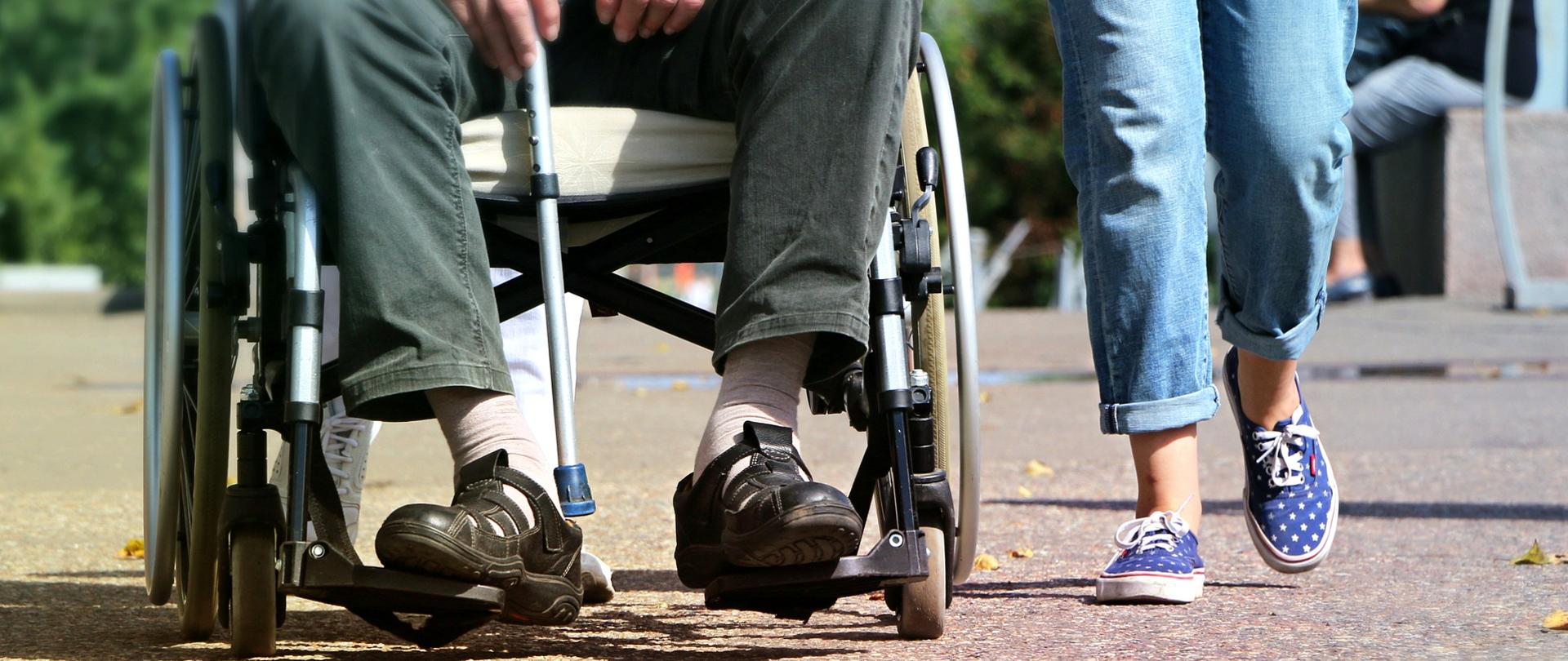 Po lewej osoba na wózku inwalidzkim, po prawej osoba sprawna idąca obok