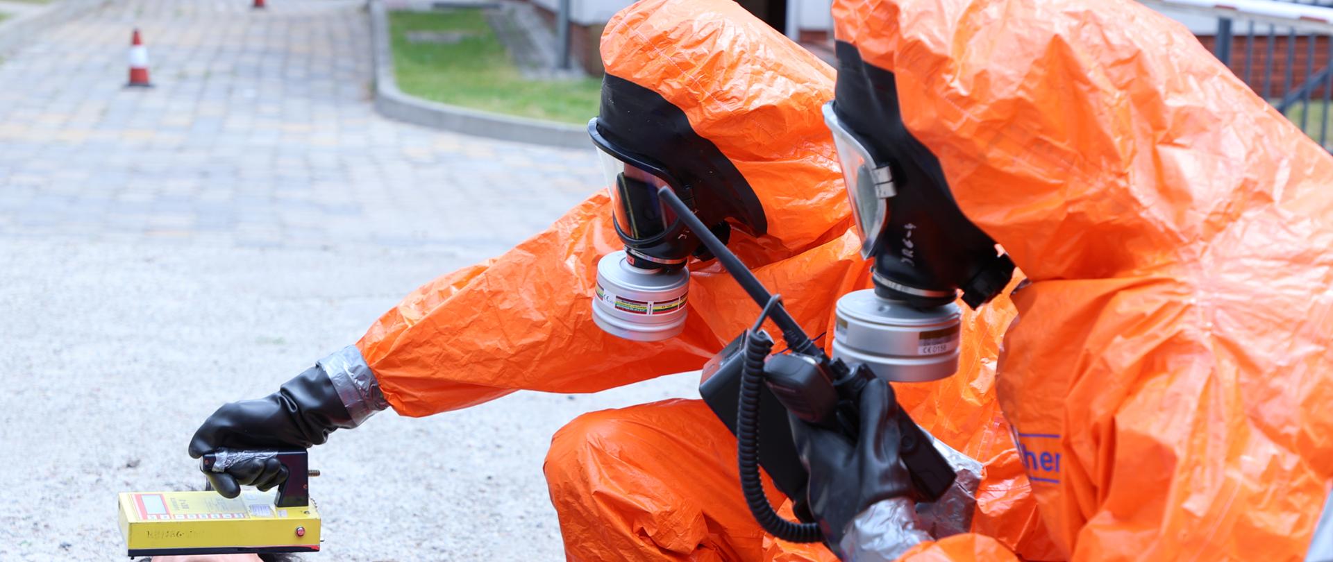 Na zdjęciu widać dwóch strażaków, w kombinezonach ochronnych w kolorze pomarańczowym z urządzeniami do pomiaru źródeł promieniotwórczych. Strażacy dokonują pomiaru przy manekinie symulującym osobę poszkodowaną.