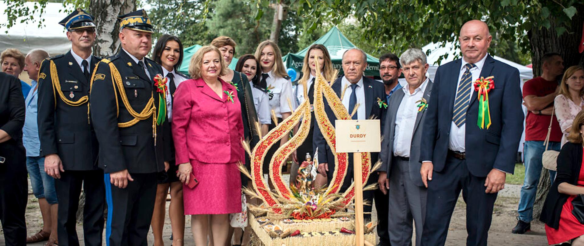 Zdjęcie wykonane na zewnątrz. Na fotografii stoją uczestnicy gminnych dożynek, wśród nich dwaj oficerowie w Państwowej Straży Pożarnej, jeden z nich w stopniu generała. W środkowej części kadru, przed uczestnikami uroczystości stoi wieniec dożynkowy. Wieniec wykonany jest ze zbóż oraz ziaren, ma kształt płomienia, w środku którego znajduje się figura Świętego Floriana. 