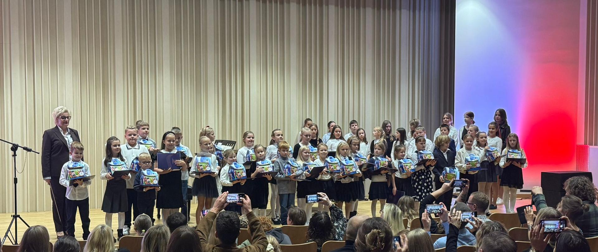 Na scenie duża grupa odświętnie ubranych dzieci, którzy stoją i trzymają dyplomy oraz słodkie upominki. W tle za nimi kobieta blondynka w okularach i czarnym garniturze, która trzyma smyczek skrzypcowy. Tło obrazka w odcieniach beżu. Zdjęcie zostało wykonane podczas koncertu z okazji Dnia Edukacji Narodowej połączonego z uroczystym ślubowaniem uczniów klas pierwszych. U samegoi dołu obrazka licznie zgromadzona publiczność.