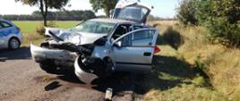 Samochód osobowy stoi na drodze po wypadku. Cała przednia część uszkodzona. Z boku stoi radiowóz policyjny. 