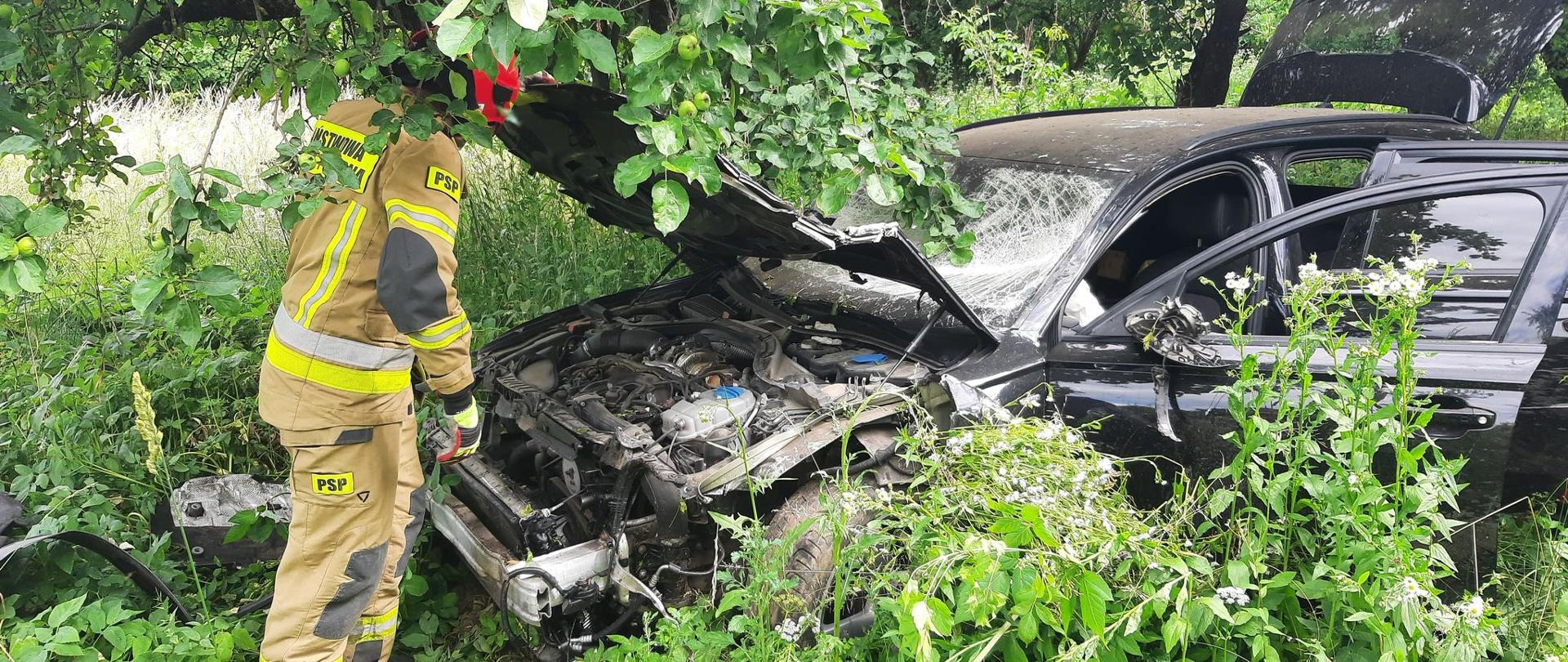 Zdjęcie przedstawia uszkodzony pojazd osobowy w obrębie, którego strażacy prowadzą działania