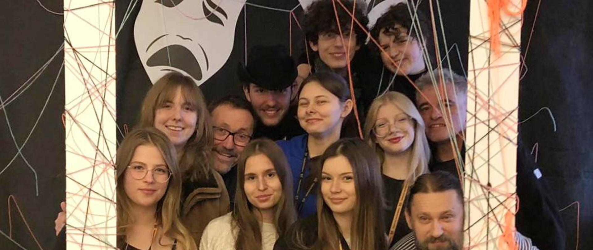 Na czarnym tle stoi dwanaście osób: 3 mężczyzn, dwóch chłopców i siedem dziewczyn. Głowy trzymają w dużej białej ramie oplecionej gęsto kolorowymi nićmi. Nad nimi białe symbole masek teatralnych. Na górze ramy logotyp: kółko z symbolem skrzydeł w środku i napis sokolowska_kultura. Na dolnej ramie symbol serca, dymku dialogowego i strzałki, cztery kropki i napis : Szpulka nawijamy teatralnie!