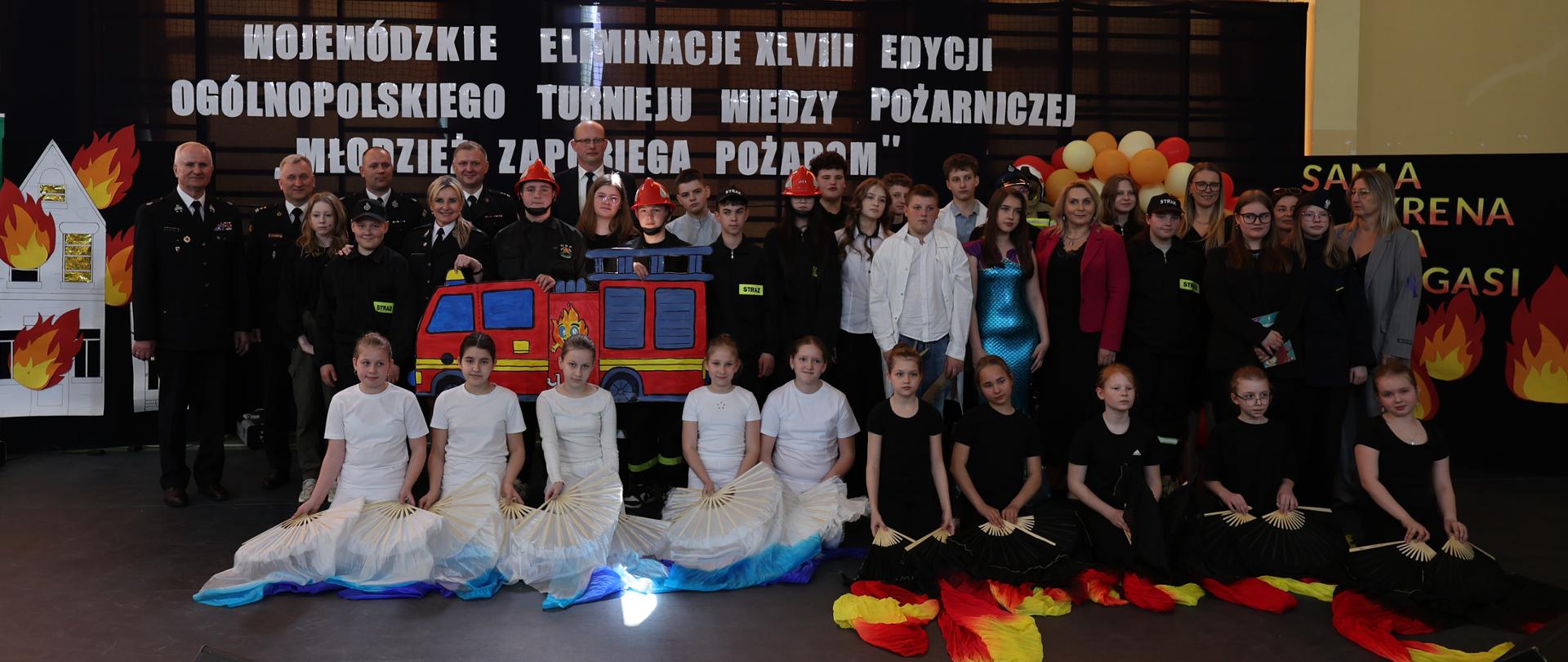 Dzieci występujące artystycznie pozują do zdjęcia przed ciemną ścianką z białym napisem „WOJEWÓDZKIE ELIMINACJE XLVIII EDYCJI OGÓLNOPOLSKIEGO TURNIEJU WIEDZY POŻARNICZEJ ‘MŁODZIEŻ ZAPOBIEGA POŻAROM’”.