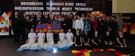 Dzieci występujące artystycznie pozują do zdjęcia przed ciemną ścianką z białym napisem „WOJEWÓDZKIE ELIMINACJE XLVIII EDYCJI OGÓLNOPOLSKIEGO TURNIEJU WIEDZY POŻARNICZEJ ‘MŁODZIEŻ ZAPOBIEGA POŻAROM’”.
