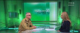Na zdjęciu przedstawiono rozmowę prowadzoną w studiu telewizyjnym TVP3 Wrocław. Przy szklanym stole siedzą naprzeciw siebie funkcjonariusz Państwowej Straży Pożarnej oraz dziennikarka prowadząca program. W tle widoczna jest scenografia programu „Rozmowa Dnia Fakty”, utrzymana w zielonej kolorystyce. Fotografia została wykonana podczas wywiadu dotyczącego zagadnień związanych z bezpieczeństwem, działalnością PSP lub bieżącymi wydarzeniami.