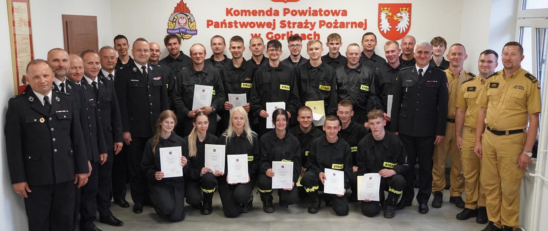 Na zdjęciu strażacy z Ochotniczych Straży Pożarnych ubrani w ubrania koszarowe koloru czarnego, zastępca komendanta powiatowego Państwowej Straży Pożarnej w Gorlicach i Prezes Zarządu Oddziału Powiatowego Związku Ochotniczych Straży Pożarnych Rzeczypospolitej Polskiej w Gorlicach oraz komisja egzaminacyjna ubrani w ubrania służbowe koloru piaskowego po egzaminie kończącym szkolenie podstawowe strażaków ratowników OSP w sali konferencyjnej w Komendzie Powiatowej Państwowej Straży Pożarnej w Gorlicach.