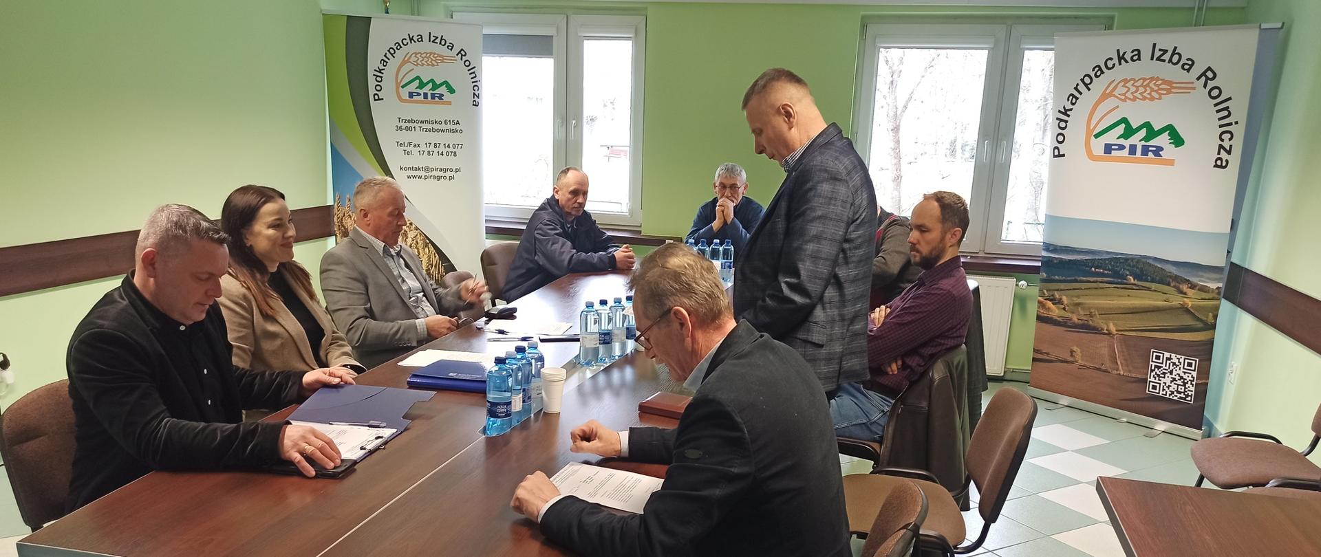 Grupa około dziesięciu dorosłych osób siedzi przy prostokątnym stole konferencyjnym w jasnozielonej sali. Uczestnicy spotkania są ubrani w stylu biznesowym – marynarki, koszule i swetry. Na stole stoją butelki wody, dokumenty oraz teczki. Jedna osoba stoi przy stole i czyta lub omawia dokument, podczas gdy pozostali słuchają lub przeglądają materiały. W tle widoczne są dwa roll-upy z napisem „Podkarpacka Izba Rolnicza” oraz okna wpuszczające światło dzienne. Atmosfera jest formalna i skupiona.