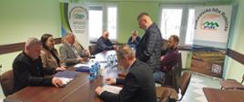 Grupa około dziesięciu dorosłych osób siedzi przy prostokątnym stole konferencyjnym w jasnozielonej sali. Uczestnicy spotkania są ubrani w stylu biznesowym – marynarki, koszule i swetry. Na stole stoją butelki wody, dokumenty oraz teczki. Jedna osoba stoi przy stole i czyta lub omawia dokument, podczas gdy pozostali słuchają lub przeglądają materiały. W tle widoczne są dwa roll-upy z napisem „Podkarpacka Izba Rolnicza” oraz okna wpuszczające światło dzienne. Atmosfera jest formalna i skupiona.