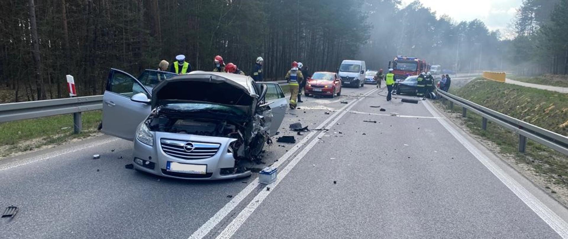Zdjęcie przedstawia na pierwszym planie przód rozbitego pojazdu osobowego. Na drugim planie znajdują się strażacy prowadzący medyczne czynności ratownicze oraz zabezpieczający teren działań. Na jezdni zaparkowane są także inne pojazdy straży, policji oraz drugi uszkodzony samochód osobowy, biorący udział w zdarzeniu. Droga wojewódzka, na której doszło do zdarzenia znajduje przebiega wzdłuż lasu. 