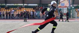 zawodnik XII powiatowych zawodów sportowo-pożarniczych Ochotniczych Straży Pożarnych z terenu powiatu łowickiego biegnie po murawie boiska z wężem pożarniczym w ręku, ubrany w ubranie koszarowe barwy czarnej z hełmem barwy białej na głowie