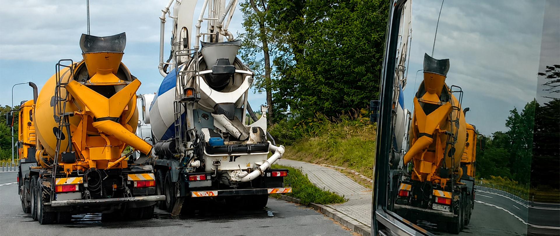 Dwa samochody ciężarowe - betonomieszarki - stoją przy drodze. Po prawej pojazd ITD. Żółta betonomieszarka jest widoczna w odbiciu szyby pojazdu.