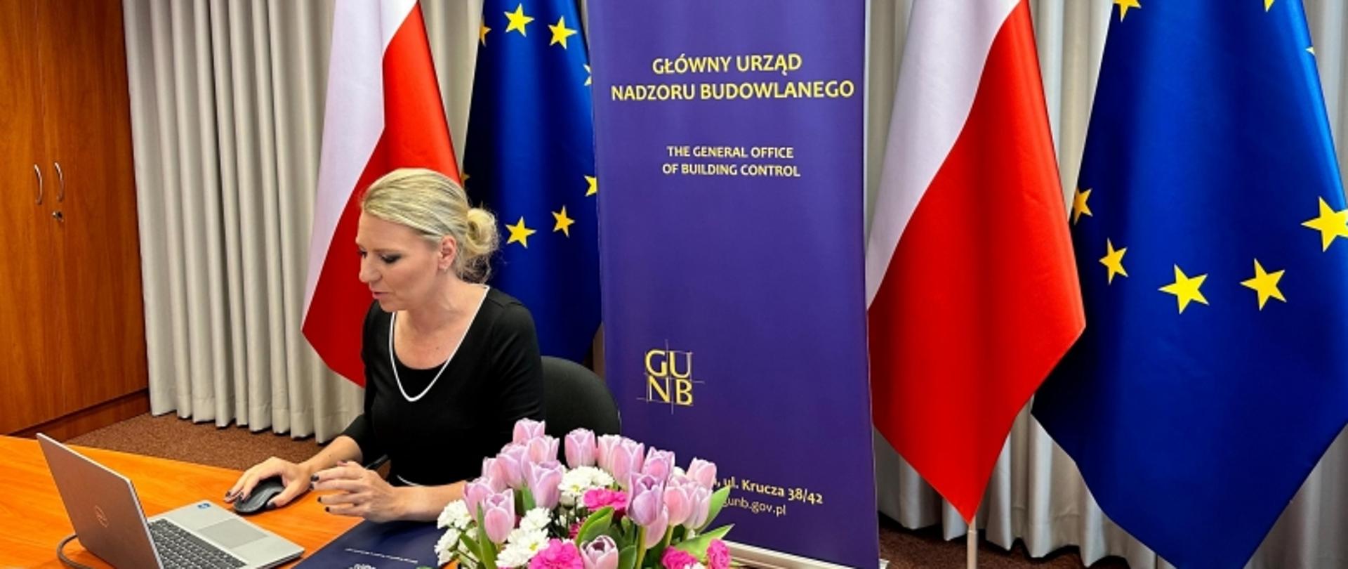 „Osoba siedząca przy biurku z laptopem, za nią baner GUNB oraz flagi Polski i UE, na biurku bukiet różowych kwiatów.”