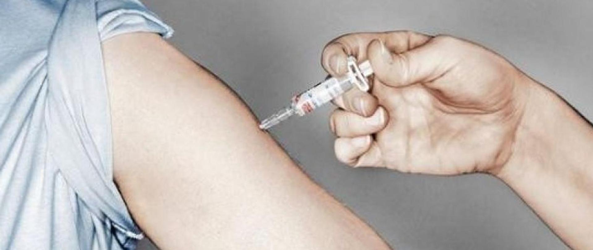 W ramię wbita jest igła ze strzykawką ze szczepionką.