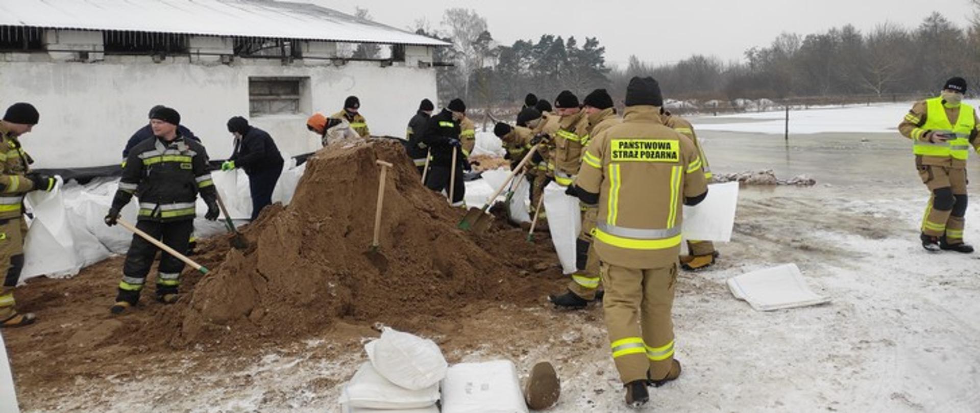 Zdjęcie przedstawia strażaków napełniających worki piaskiem, w tle budynek gospodarczy.