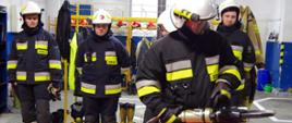 Widok z przodu. Na pierwszym planie strażak OSP w ubraniu specjalnym i hełmie trzyma w rękach nożyce hydrauliczne. W tle stoi w szeregu czterech strażaków OSP w ubraniach specjalnych i hełmach.