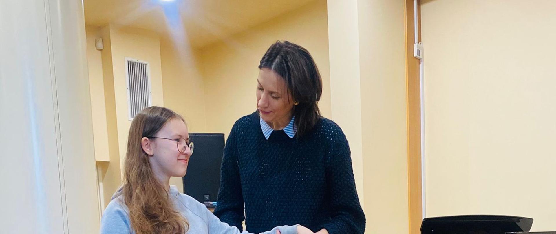 Na scenie stoi uczennica która ma wyciągniętą przed siebie rękę. Za rękę trzyma uczennicę pani która prowadzi warsztaty. Po prawej stronie stoi fortepian a na ziemi leżą 2 wiolonczele