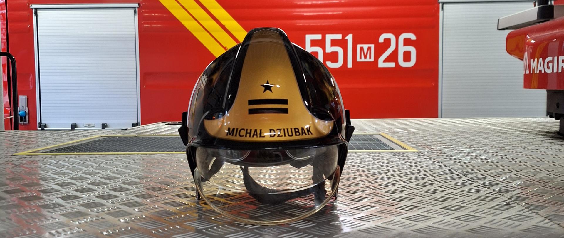 Na zdjęciu widać strażacki hełm leżący na metalowej, ryflowanej podłodze wewnątrz remizy.
Co dokładnie przedstawia fotografia
• Hełm jest złoto czarny, z przezroczystą przyłbicą.
• Na jego powierzchni widnieje imię i nazwisko „MICHAL DZIUBAK”, a obok czarna gwiazda i dwa poziome pasy.
• W tle stoi czerwony wóz strażacki z żółtymi pasami ostrzegawczymi i numerem operacyjnym 551[M]26.
• Widać fragmenty wnętrza strażnicy — oświetlenie, konstrukcję sufitu i podłogę garażową.

