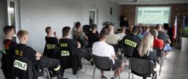 grupa kilkunastu słuchaczy siedzi na krzesłach w pięciu rzędach na sali, przed nimi zastępca komendanta powiatowego ubrany w ubranie sztabowe stoi przed stolikiem, trzyma w dłoni zwiniętą kartkę, drugi strażak ubrany w ubranie sztabowe siedzi za stolikiem, za ich plecami ściana z cegły i ekran projekcyjny na którym wyświetla się strona internetowa z zagadnieniami szkoleniowymi strażaków osp, po ich prawej stronie stoi duża donica z rośliną o drobnych liściach, na ścianie za nimi po bokach ekranu widoczny herb powiatu łowickiego oraz logo Państwowej Straży Pożarnej, w prawym rogu duża donica z rośliną o drobnych liściach oraz stojak a w nim trzy flagi, polski, powiatu łowickiego oraz unii europejskiej, pięć osób na sali ubrana jest w ubranie koszarowe osp