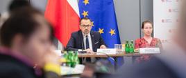 Pierwsze posiedzenie KM KPO - wiceminister funduszy i polityki regionalnej Marcin Horała siedzi przy stole i przemawia przez mikrofon, za jego plecami flagi Polski i UE 