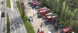 Widok z lotu ptaka na zgrupowanie służb ratowniczych. Wzdłuż drogi zaparkowanych jest kilkanaście pojazdów: czerwone wozy strażackie, białe karetki pogotowia oraz radiowozy policji. Przy pojazdach widać grupy ratowników.