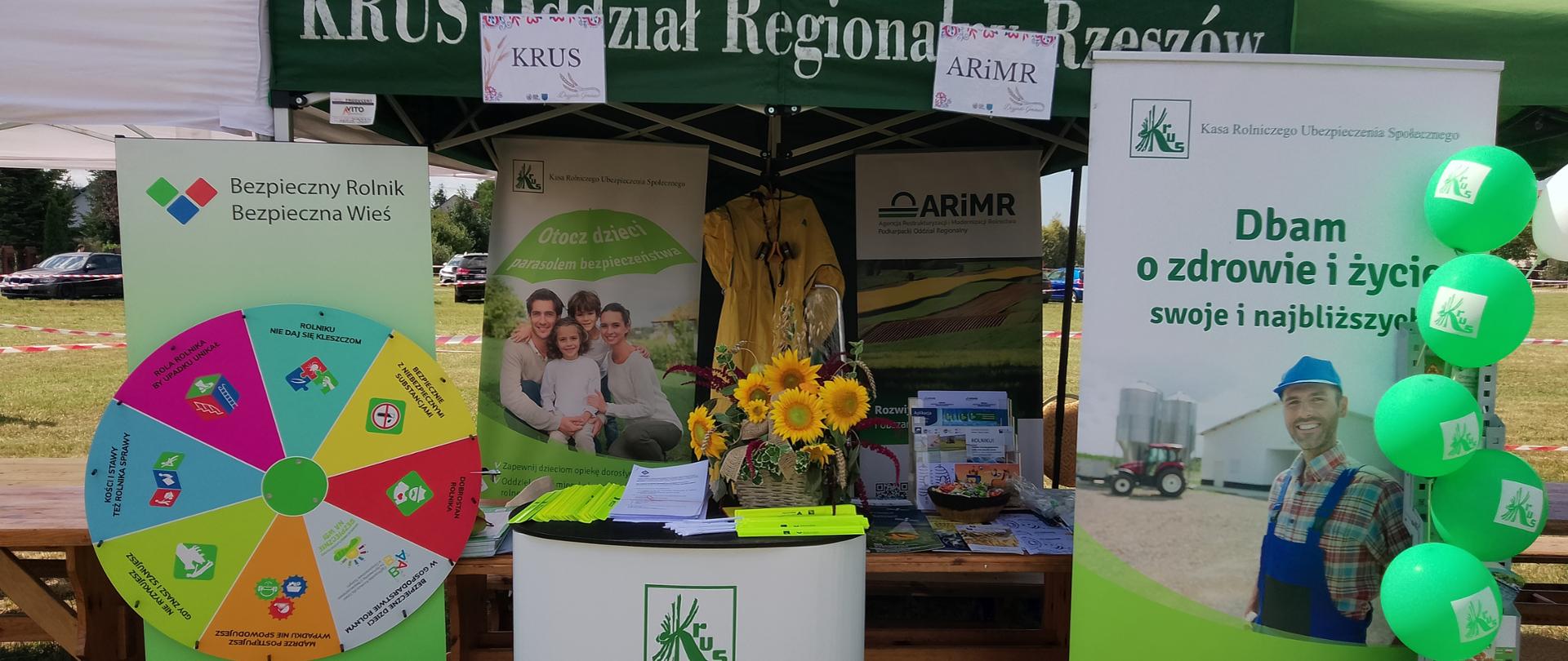 Na tle banerów z logiem KRUS oraz napisem kampanii prewencyjnych KRUS znajduje się stolik, na którym leżą ulotki oraz materiały prewencyjne Kasy.